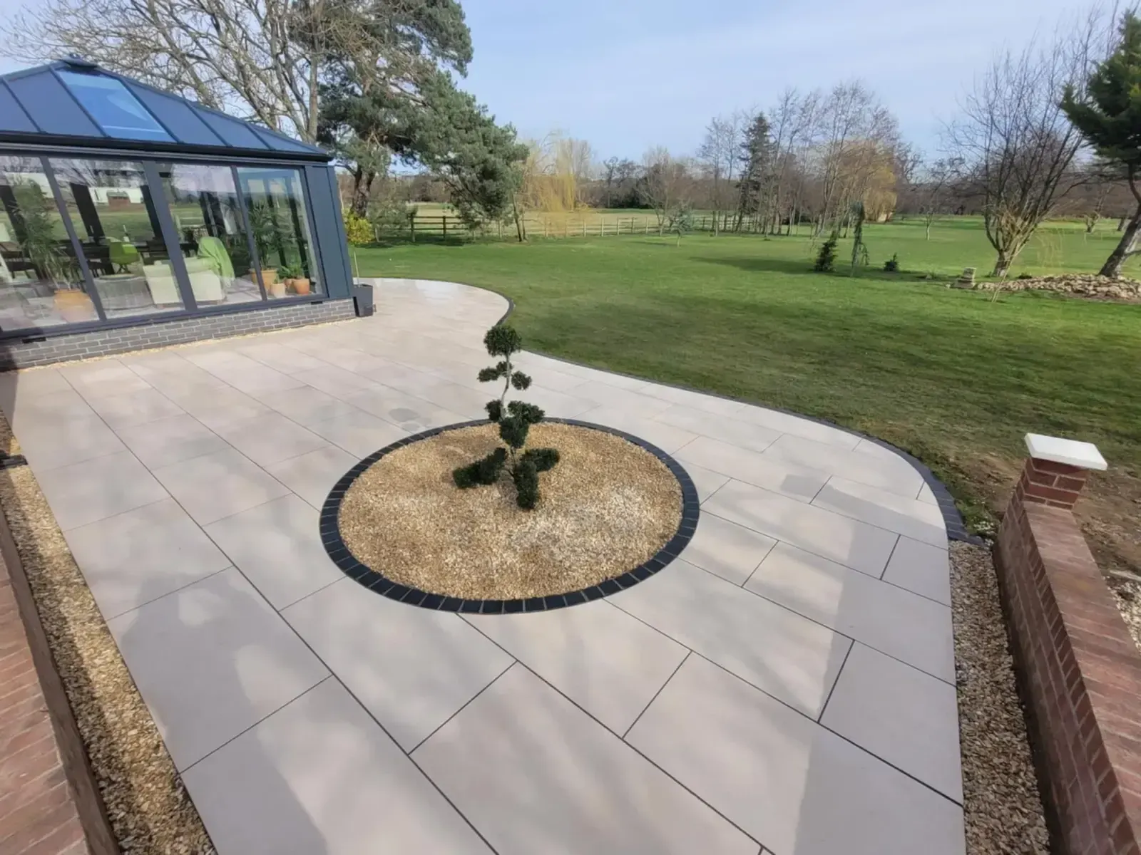 Curved porcelain patio around a circular planting bed by an orangery