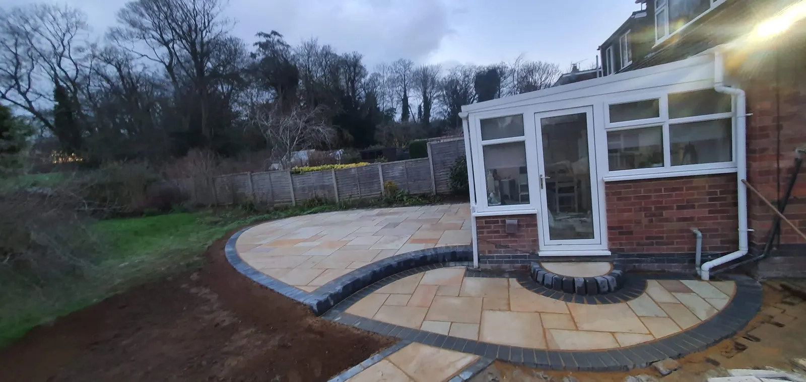 Curved sandstone patio with a charcoal border round a conservatory at dusk