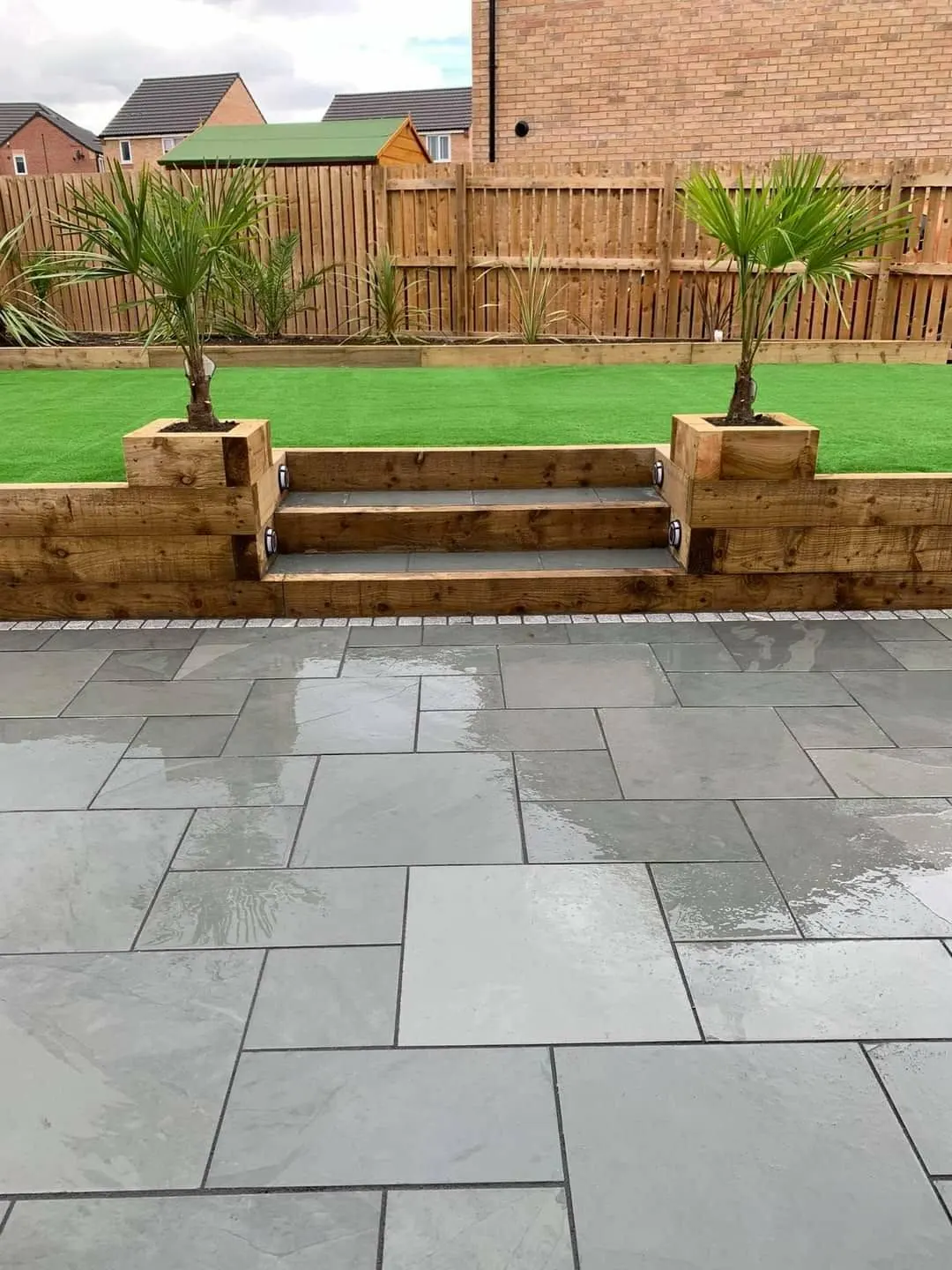 Sleeper retaining wall with steps and palm planters above a grey porcelain patio