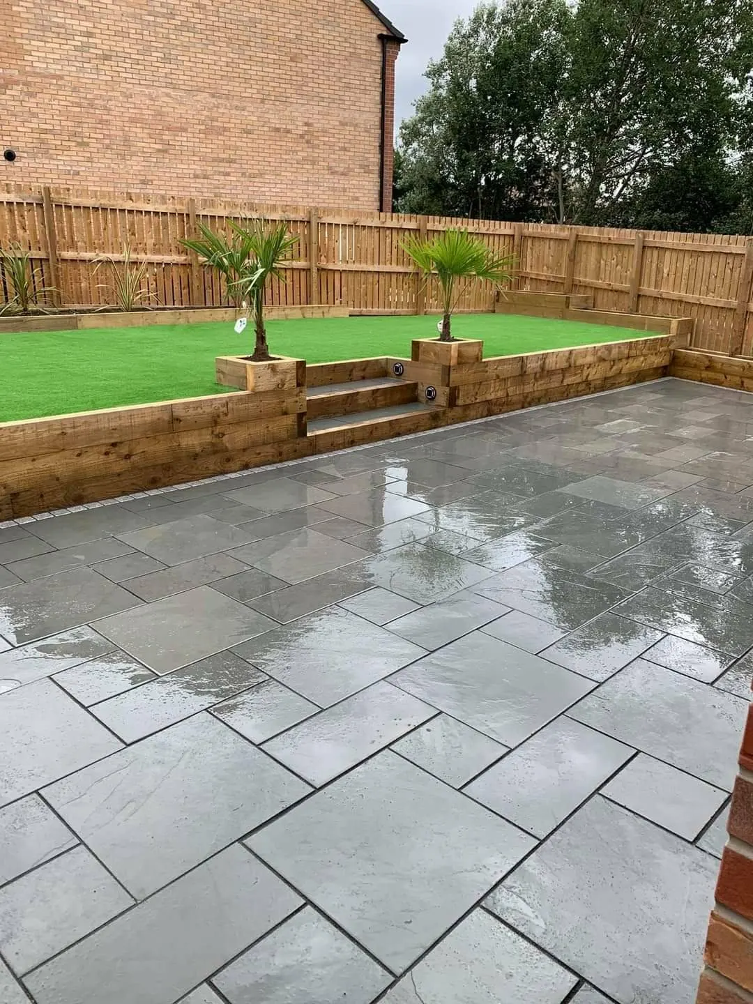Grey porcelain patio after rain below a sleeper retaining wall and artificial grass
